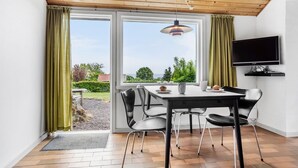 Un espace repas comprenant une table et des chaises, un téléviseur fixé au mur et une vue sur l’extérieur grâce à de grandes fenêtres.