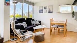 Een moderne woonkamer met een zwarte bank, een witte salontafel en een houten eettafel met stoelen. Grote ramen zorgen voor natuurlijk licht en uitzicht op groen buiten.