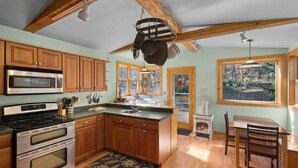 A kitchen with wooden cabinets, a microwave, stove, and a large window with a view of trees. - A-Frame Home,2mi from Mt.Snow w/Wood Burning Stove (Dover)