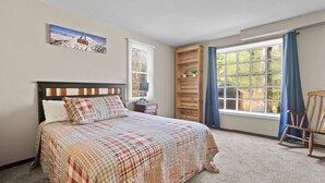 A bedroom with a bed, a nightstand, a wooden shelf, a rocking chair, a window with curtains, and a painting on the wall. - A-Frame Home,2mi from Mt.Snow w/Wood Burning Stove (Dover)