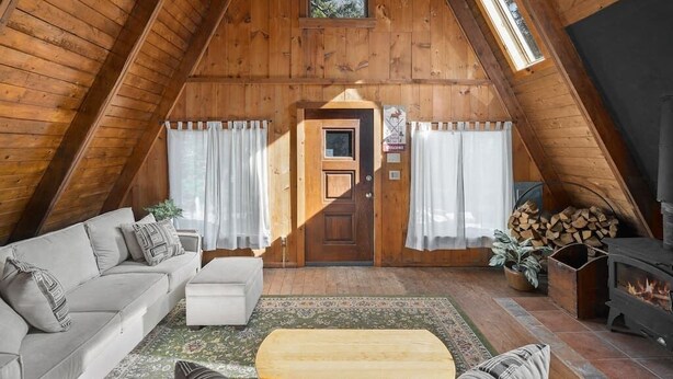 A cozy attic room with a wooden ceiling, a sofa, a small table, a fireplace, and a door with curtains. - A-Frame Home,2mi from Mt.Snow w/Wood Burning Stove (Dover)