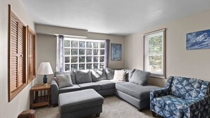A living room with a sectional sofa, an ottoman, a patterned armchair, a lamp, and a window with curtains. - A-Frame Home,2mi from Mt.Snow w/Wood Burning Stove (Dover)