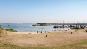 Uma praia com um porto de iates ao fundo, areal arenoso e pessoas relaxando.