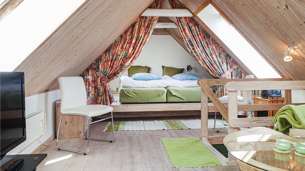 Um quarto de sótão acolhedor, com teto inclinado, cama com roupa de cama verde, uma cadeira branca, uma mesinha com velas e uma televisão.