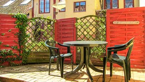 Um pátio externo com mesa e duas cadeiras, cerca vermelha e um edifício amarelo com janelas.