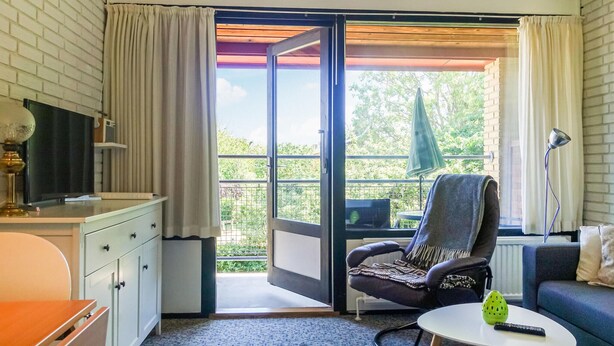 A room with a desk, a chair, a small table, and a sofa. There is a sliding glass door leading to an outdoor area with a view of greenery.