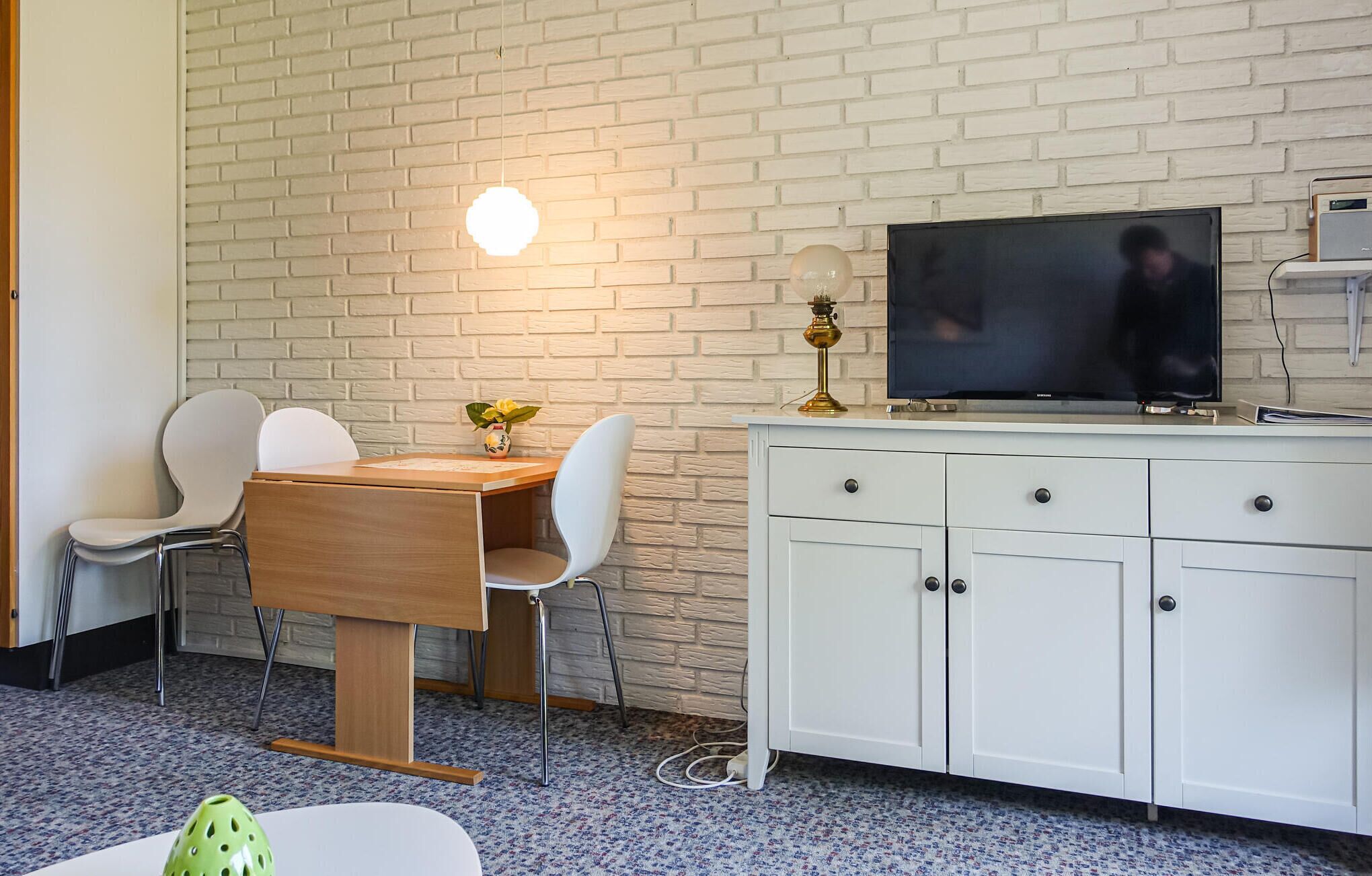 Quarto moderno com parede de tijolos brancos, mesa de jantar de madeira com duas cadeiras brancas, armário branco com gavetas e uma TV de tela plana em cima.