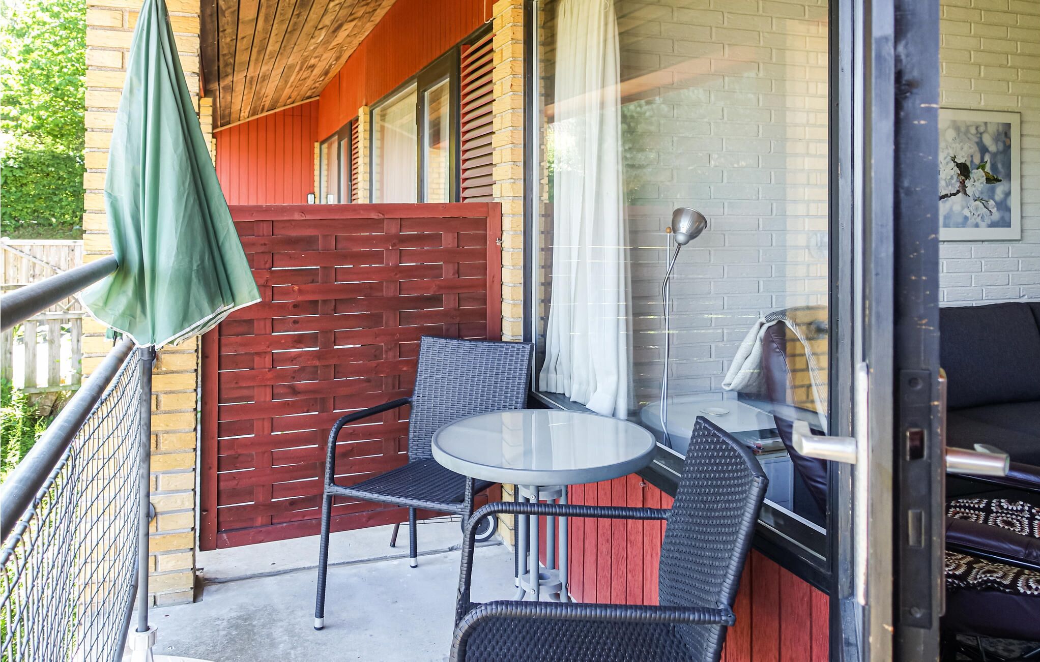 Un balcon avec des fauteuils en osier et une petite table ronde, une clôture en bois rouge et une porte vitrée donnant sur une pièce où se trouvent un canapé et une lampe.