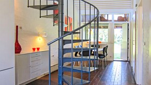 Une cuisine moderne avec un escalier en colimaçon bleu menant à un niveau supérieur. On aperçoit un coin repas avec une table et des chaises à travers une porte ouverte.