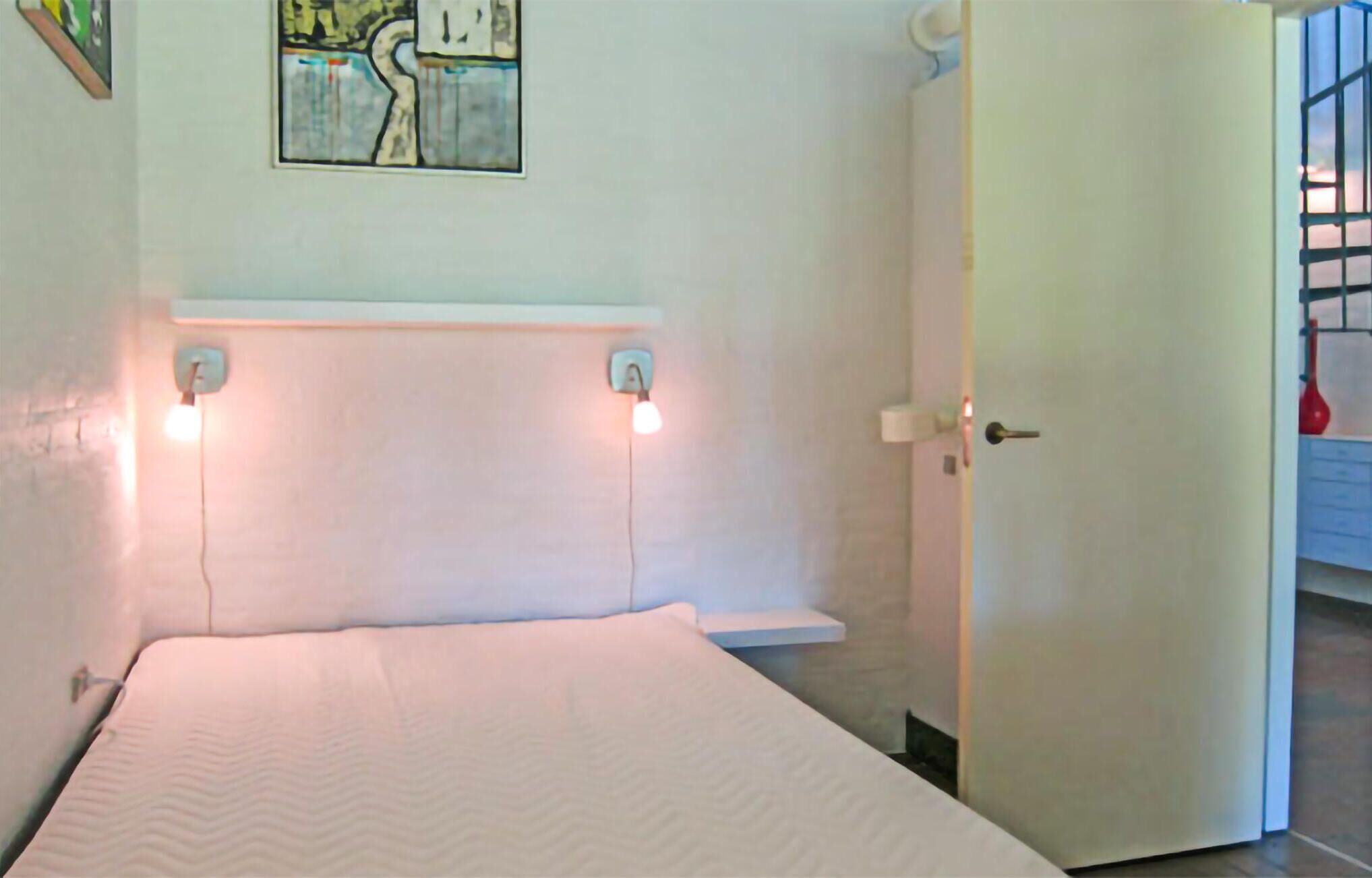 A bedroom with a bed, two bedside lamps, a painting on the wall, and a door leading to another room.
