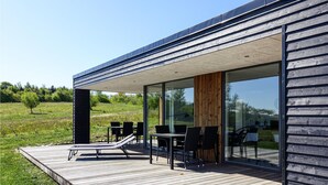 Une terrasse extérieure moderne, dotée d’un espace repas comprenant des tables et des chaises, et d’une plateforme en bois menant à une porte vitrée.