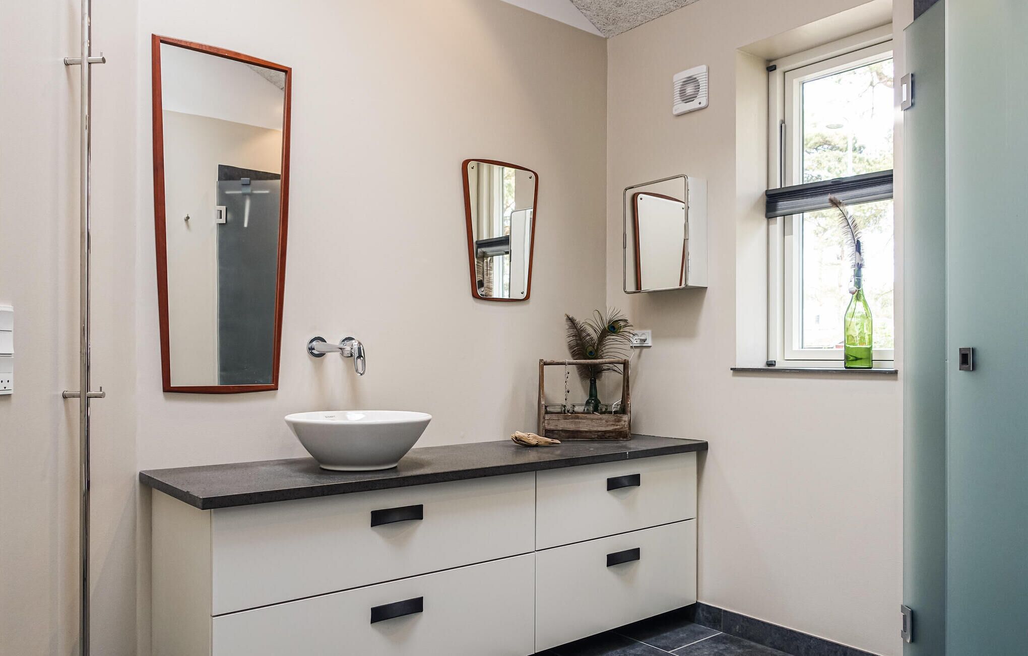 Une salle de bain avec un lavabo blanc, deux miroirs, une fenêtre et une porte.