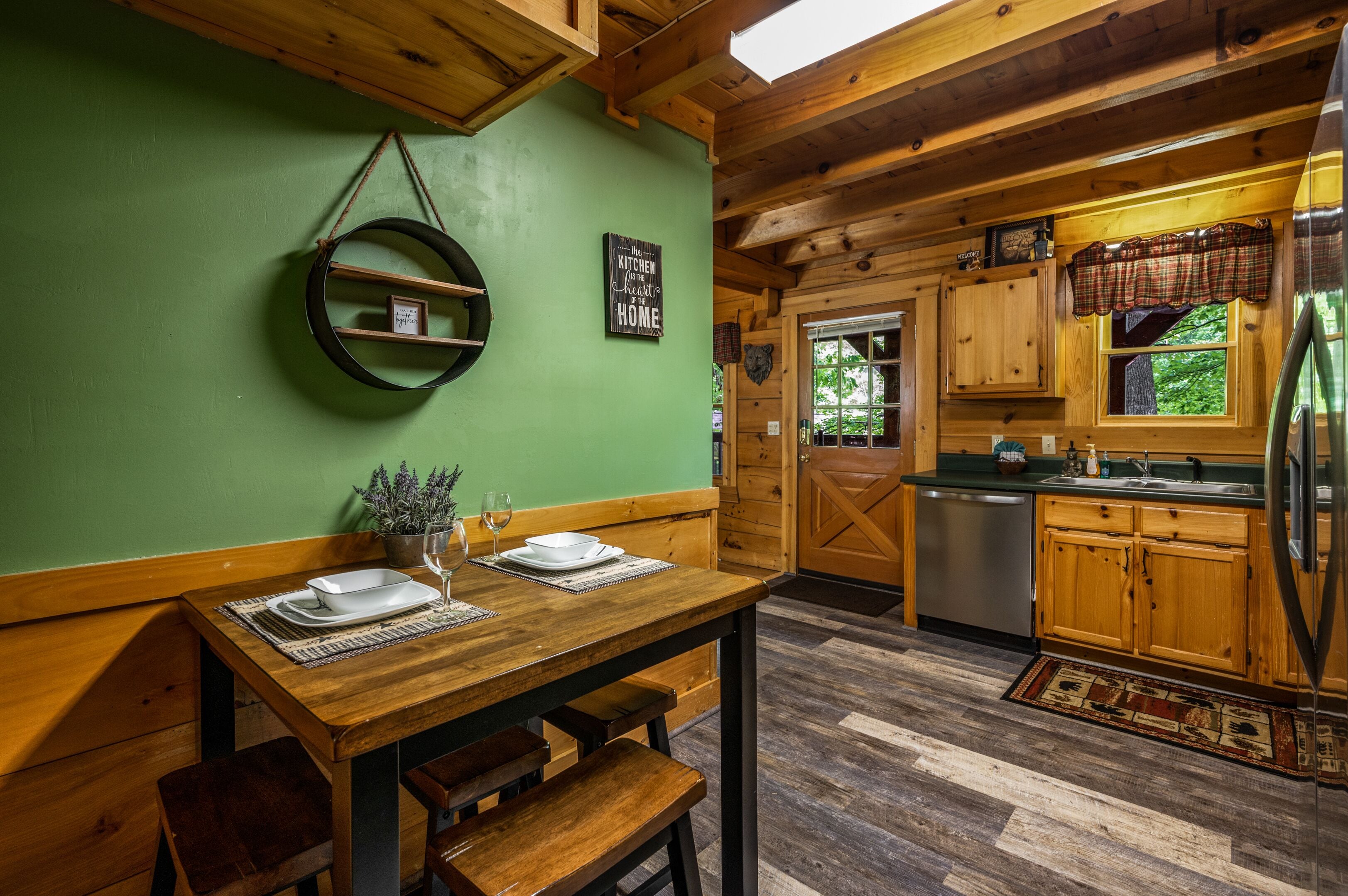 A wooden dining table set with plates and a wine glass, a wooden bench, a green wall, a kitchen with wooden cabinets and a stainless steel refrigerator.