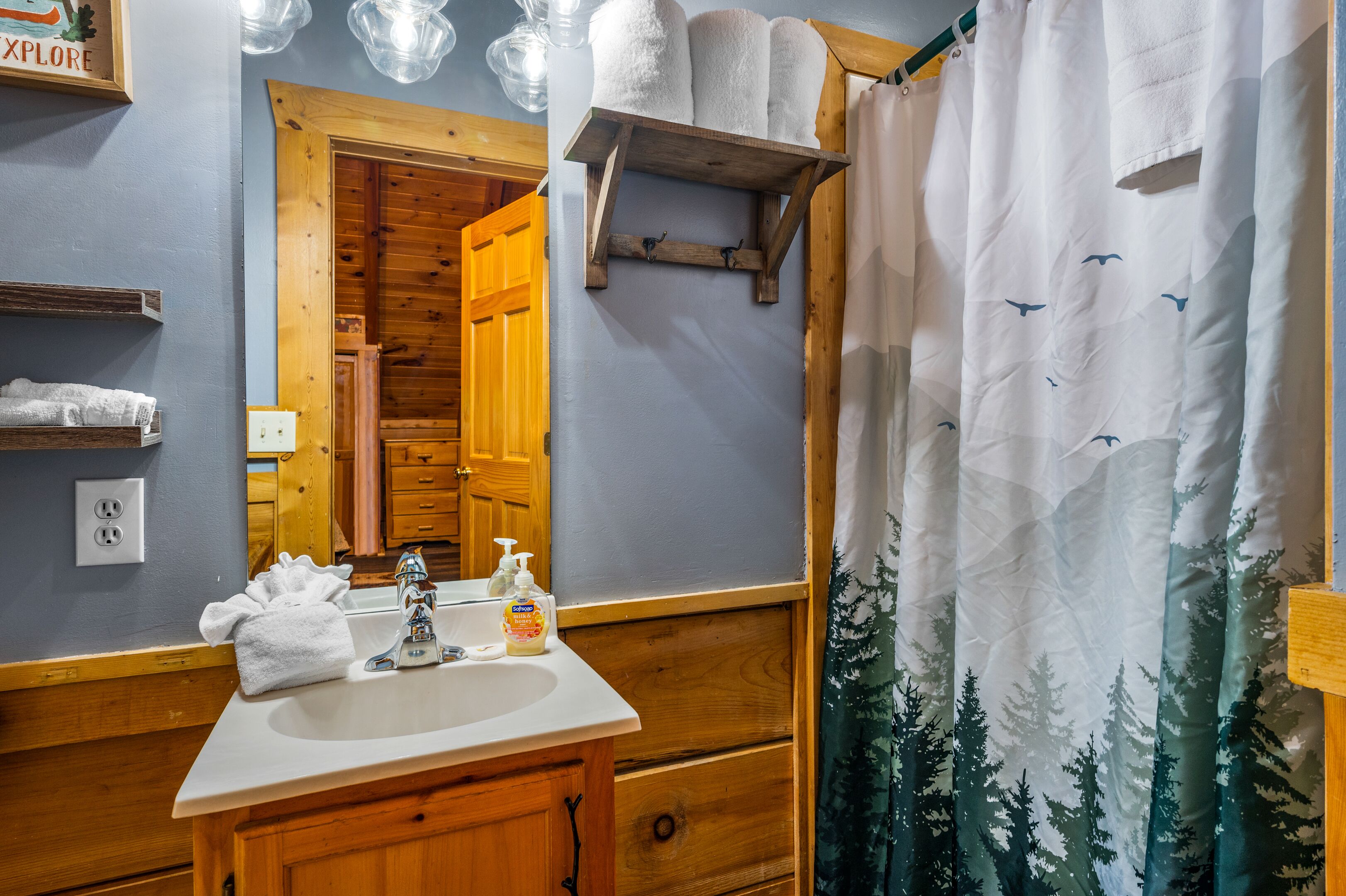 A bathroom with a wooden vanity, a sink, a mirror, and a shower curtain with a forest design.