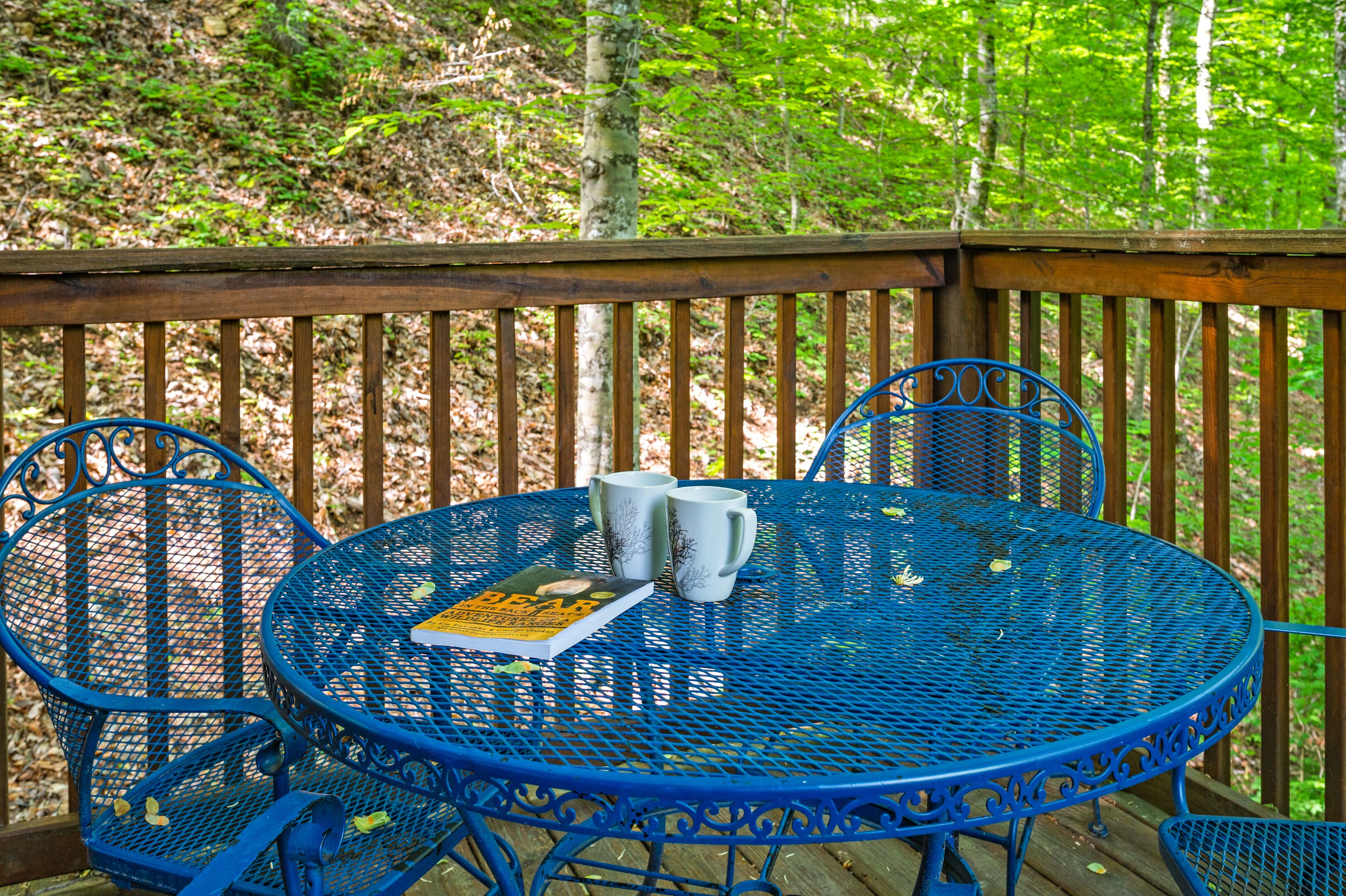 A blue metal table with two white mugs and a book on a wooden deck surrounded by trees.
