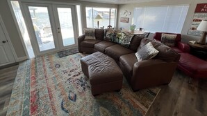 A spacious living room with a brown leather sectional sofa, a colorful area rug, and a large window with blinds.