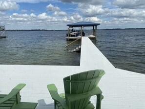 A wooden dock extending into a body of water with a covered structure at the end, and green chairs in the foreground.