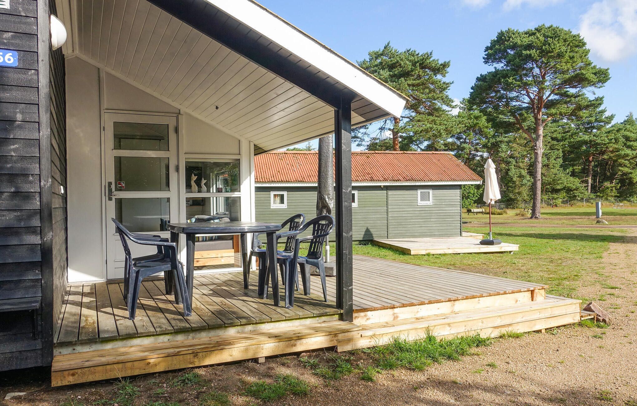 Eine Holzterrasse mit Tisch und Stühlen, eine überdachte Veranda und ein kleines Gartenhaus auf einer Rasenfläche mit Bäumen.