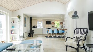 Un ampio soggiorno con soffitto alto, un'area pranzo con sedie blu, una poltrona nera con una coperta in pelliccia e una cucina sullo sfondo.