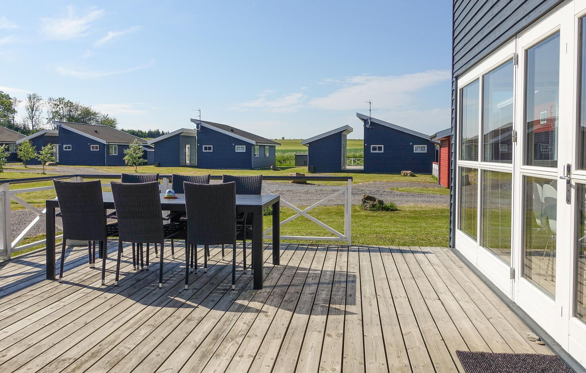 A wooden deck with a table and chairs, overlooking a row of blue buildings with a grassy field in the background.