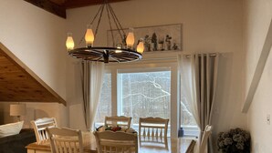 A dining area with a wooden table and chairs, a chandelier, and a view of snow-covered trees through the window. - The Barn Loft.  Streamside  living in a spacious and tastefully renovated barn (Roxbury)
