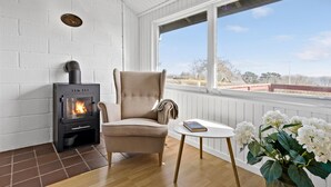 A cozy room with a wood-burning stove, a comfortable armchair, a small table with a book, and a view of the outside. - Beautiful home in Allinge with WiFi (Allinge)