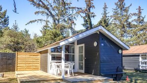 Una cabaña pequeña de color oscuro con una terraza de madera y una puerta de cristal que da a un interior con una mesa y sillas.