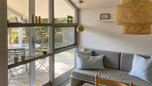 Un cuarto acogedor con un sofá gris, una silla de madera y una mesa. Hay un ventanal con vista a una terraza de madera y vegetación al exterior.