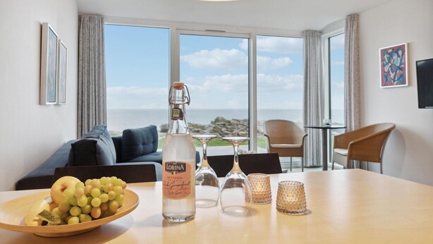 Habitación de hotel con una mesa de comedor servida con una botella de licor, vasos y un frutero. Se observa un sofá y sillas al fondo, y un ventanal con vista al mar.