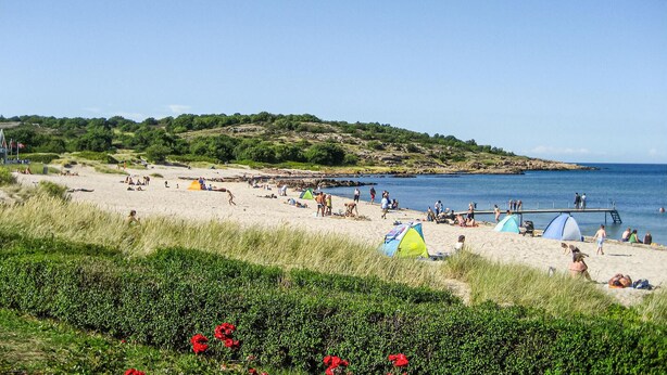 En strand med tält och människor, en brygga och ett kuperat landskap i bakgrunden.