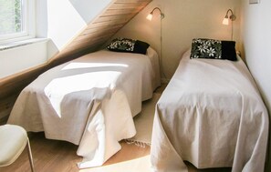 Ein Zimmer mit zwei Betten, jedes mit einem Kopfkissen und einer Nachttischlampe. Ein Fenster lässt natürliches Licht herein und es gibt einen Sessel in der Ecke.
