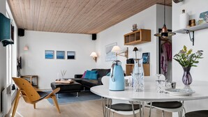 Een moderne woonkamer met een zwarte leren bank, een houten stoel, een witte eettafel met vier stoelen, en een keukenhoek met een koffiezetapparaat en een bloevas.