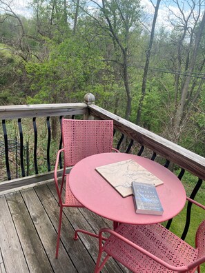 Une terrasse en bois avec une table et une chaise en m étal roses, un livre intitulé « L’univers élégant » de Brian Greene, et une vue sur une zone boisée.