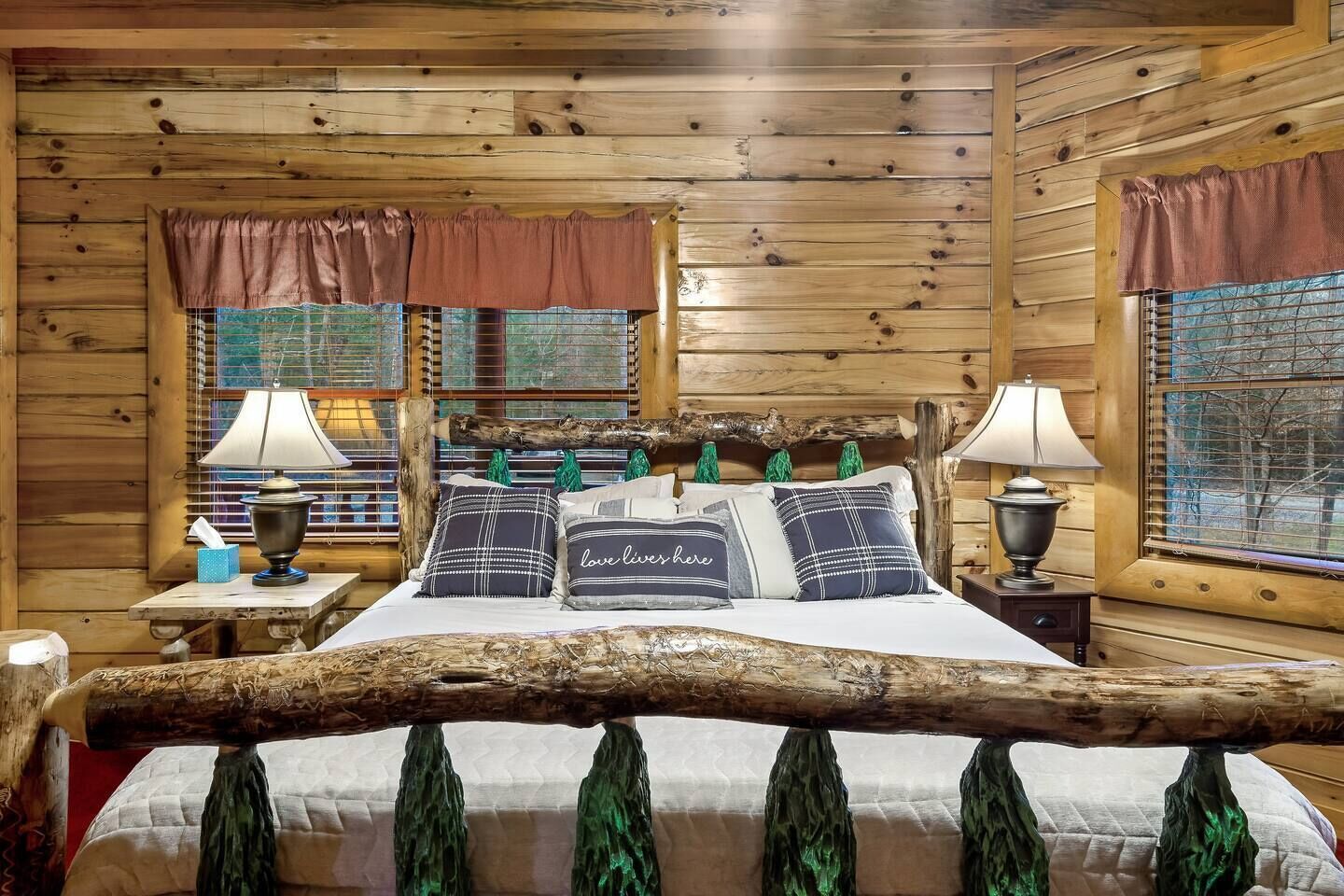 Une chambre de style cabane avec un lit placé au centre, sous une poutre en bois rustique, flanqué de deux fenêtres équipées de stores, d’une table de chevet avec une lampe et d’une petite table de nuit avec une autre lampe.