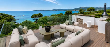 A deck with outdoor furniture, a table with wine glasses, and a view of the sea and hills.