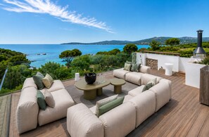 A deck with outdoor furniture, a table with wine glasses, and a view of the sea and hills.