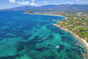 An aerial view of a coastal area with clear blue waters, boats, and a beach with buildings and greenery.