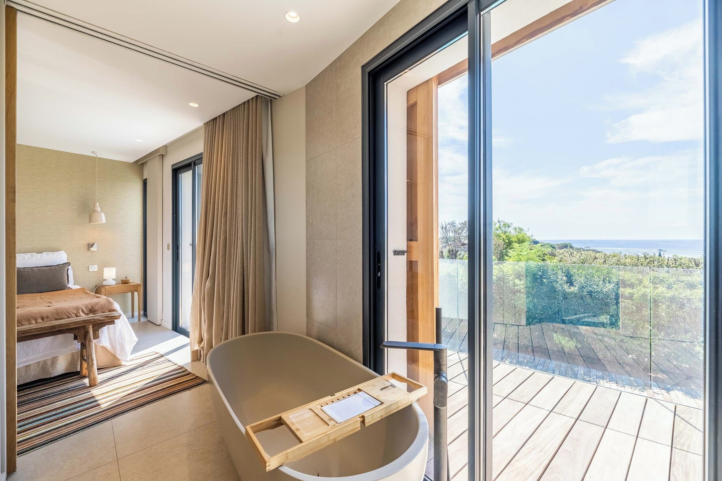 A modern bedroom with a large bed, a wooden bedside table, a bedside lamp, and a view of the outdoors through a sliding glass door.