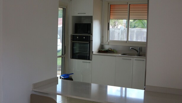 Una cocina moderna con armarios blancos, un horno empotrado y un ventanal con persianas. En primer plano, hay dos sillas y una mesa pequeña.