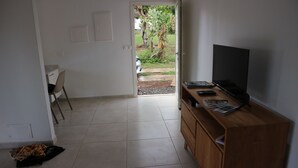 Una habitación con un mueble bajo de madera para el televisor, un televisor de pantalla plana y un pequeño escritorio con una silla. Hay una puerta que da a un área exterior con jardín y un árbol.