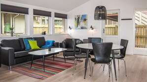 Una sala de estar moderna con un sofá negro, almohadas coloridas, un tapete a rayas, una mesa de comedor redonda con sillas y un ventanal con persianas.