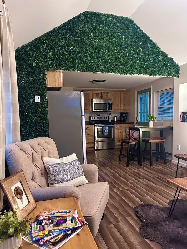 A modern living room with a sofa, a coffee table with magazines, and a kitchen area with wooden cabinets and stainless steel appliances.