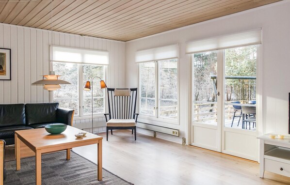 Amplia sala de estar con un sofá de cuero negro, una mesa de madera con un tazón verde y una silla a rayas. Grandes ventanales ofrecen vistas a una terraza nevada al aire libre.