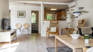 Een moderne woonkamer met houten vloer, een televisie, twee stoelen, een eettafel, en een keukenhoek met kasten en een hanglamp.