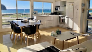 Une cuisine moderne avec des meubles blancs, un four à micro-ondes et un réfrigérateur. On y trouve un coin repas avec une table et des chaises, ainsi que de grandes fenêtres offrant une vue sur l’océan.