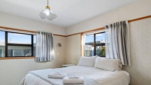Hotellihuone, jossa on suuri sänky, kaksi ikkunaa verhoilla, yöpöytä ja kattovalaisin.