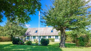 Uma casa tradicional com telhado inclinado, rodeada por um gramado bem cuidado e árvores frondosas.