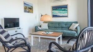 Een woonkamer met een groene bank, twee zwarte rieten fauteuils, een houten salontafel, een televisie en twee schilderijen aan de muur.