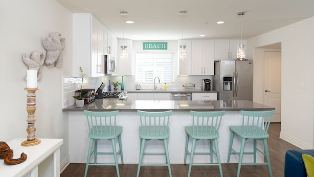 A modern kitchen with white cabinets, a central island with bar stools, and a 'BEACH' sign above the window. - 326 San Luis (Pismo Beach)