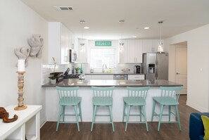 A modern kitchen with white cabinets, a central island with bar stools, and a 'BEACH' sign above the window. - 326 San Luis (Pismo Beach)